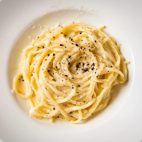 Steaming spaghetti cacio e pepe glistens in a skillet, flecked with black pepper and topped with shaved Pecorino Romano.